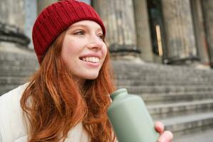 Smiling redhead girl in red hat, sits on stairs near building with flask, drinks water and looks happy photo