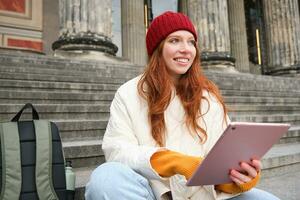 hermosa joven moderno niña con rojo cabello, sostiene digital tableta, se sienta en escalera cerca museo y conecta público Internet, envía mensaje en artilugio aplicación foto