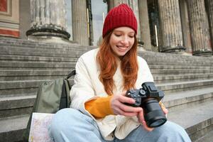 Young student, photographer sits on street stairs and checks her shots on professional camera, taking photos outdoors