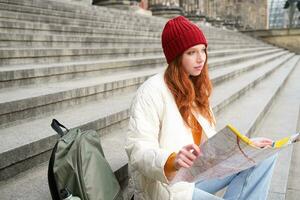 Beautiful young woman, traveler with backpack holds paper map, explores city sightseeing, plans route for tourism popular attractions photo