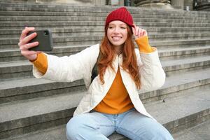 elegante joven niña en rojo sombrero, toma fotos en teléfono inteligente cámara, hace selfie como ella se sienta en escalera cerca museo, posando para foto con aplicación filtrar
