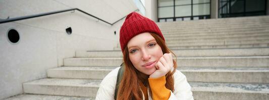 Close up portrait of beautiful redhead girl in red hat, urban woman with freckles and ginger hair, sits on stairs on street, smiles and looks gorgeous photo