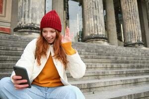 Young redhead woman sits on stairs outdoors and waves hand at smartphone camera, video chats with friends, connects to public wifi photo