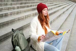 Tourism and lifestyle concept. Young redhead woman looking at city map, plans a route for sightseeing day, sits outdoors on stairs and rests photo