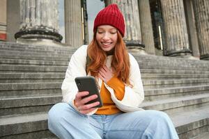 Young smiling woman with long red hair, sits near building on stairs with smartphone, using mobile application, sending message photo