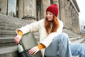 Smiling tourist, girl sits on stairs, rests on staircase, takes thermos from backpack, drinks hot coffee from flask photo