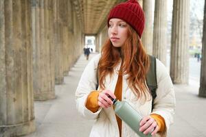 Young redhead girl holds thermos in hands, pours herself a hot drink while walking in city. Tourist takes break, opens flask for refreshment beverage photo