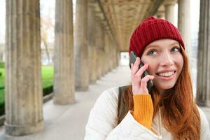 sonriente linda pelirrojo mujer hace un teléfono llamar, sostiene teléfono cerca año, tiene móvil conversación, utilizando teléfono inteligente en calle foto