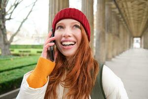 retrato de pelirrojo europeo niña en rojo sombrero, hace un teléfono llamar, camina en ciudad y negociaciones a amigo en teléfono inteligente foto