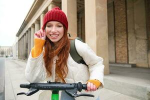 Smiling redhead woman stands near building, rents escooter to travel around town while on tourist trip photo