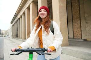 joven sonriente pelirrojo chica, estudiante paseos eléctrico scooter, alquila eso y viajes alrededor ciudad foto