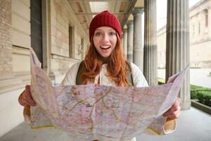 aventurero pelirrojo niña camina en pueblo con papel mapa, explora ciudad como turista, mira para popular turismo atracciones, mira alrededor emocionado y sonrisas foto