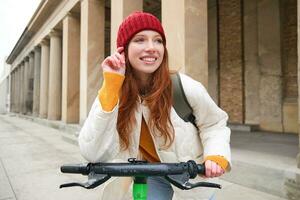 joven sonriente pelirrojo chica, estudiante paseos eléctrico scooter, alquila eso y viajes alrededor ciudad foto