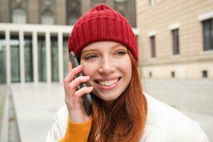 móvil conexión y personas concepto. contento pelirrojo mujer en sombrero, negociaciones en móvil teléfono, haciendo teléfono llamar, utilizando aplicación a llamada extranjero foto