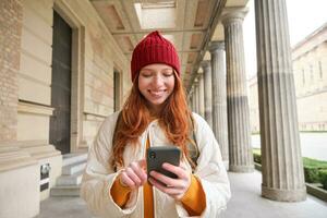 Smiling woman with red hair, tourist using mobile phone application while walking around town, searching for attraction to visit, exploring sightseeing with smartphone app photo