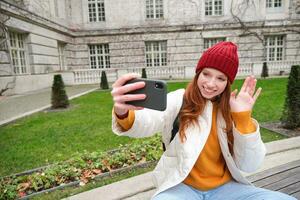 Portrait of girl blogger records video and says hi at phone camera. Young woman takes selfie with smartphone app, waves hand, video chats in application photo