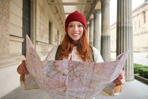 sonriente joven pelirrojo mujer en rojo sombrero, mira a papel mapa a Mira para turista atracción. turismo y personas concepto. niña explora ciudad, intentó a encontrar camino foto