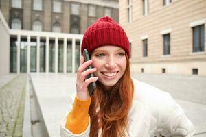 elegante moderno pelirrojo niña negociaciones en móvil teléfono, hace un teléfono llamar, vocación alguien en teléfono inteligente aplicación desde afuera, soportes en calle y sonrisas foto