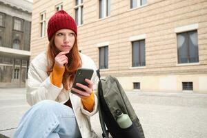 Young redhead woman with smartphone, sitting outdoors with backpack, thinking, looking thoughtful, making decision, looking at map and plans route photo