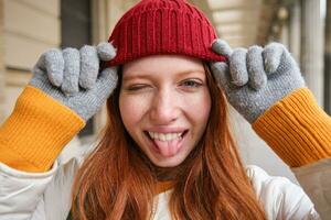 retrato de joven pelirrojo mujer en de punto sombrero y guantes, sonrisas y mira aparte, camina alrededor ciudad en invierno foto