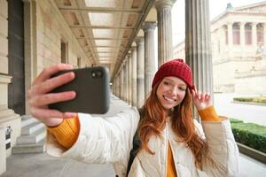 Stylish redhead girl tourist, takes selfie in front of tourism attraction, makes photo with smartphone, looks at mobile camera and poses