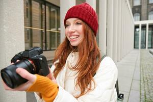 Smiling redhead girl photographer, taking pictures in city, makes photos outdoors on professional camera