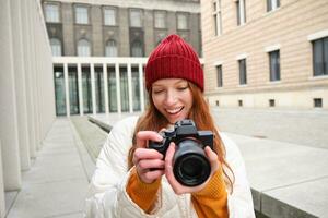 Smiling redhead girl photographer, taking pictures in city, makes photos outdoors on professional camera