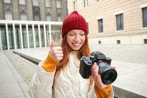 sonriente pelirrojo niña fotógrafo, tomando imágenes en ciudad, hace fotos al aire libre en profesional cámara