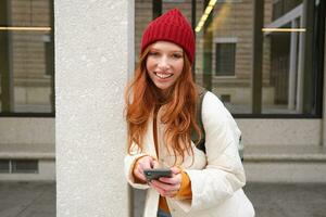 elegante urbano niña utilizando móvil teléfono aplicación, en pie en ciudad, esperando para Taxi, mirando a teléfono inteligente solicitud, mensajes de texto mensaje foto
