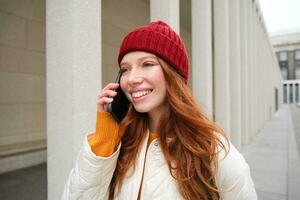 contento pelirrojo mujer, niña con teléfono negociaciones, tiene conversacion en móvil aplicación, usos Internet a llamada extranjero con teléfono inteligente aplicación, riendo y sonriente foto