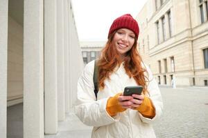 Beautiful smiling girl, tourist with backpack, holding smartphone, using map on mobile phone application, looking for sightseeing in internet app, standing outdoors photo