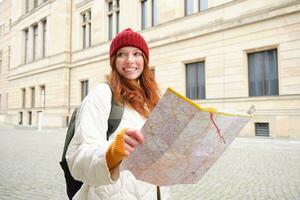 pelirrojo chica, turista explora ciudad, mira a papel mapa a encontrar camino para histórico puntos de referencia, mujer en su viaje alrededor Europa búsquedas para Turismo foto