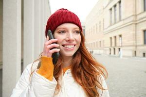 contento pelirrojo mujer, niña con teléfono negociaciones, tiene conversacion en móvil aplicación, usos Internet a llamada extranjero con teléfono inteligente aplicación, riendo y sonriente foto