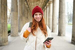 Happy young female model stands on street and holds mobile phone, uses smartphone app, follows the route of online map photo