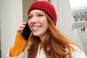 contento pelirrojo mujer, niña con teléfono negociaciones, tiene conversacion en móvil aplicación, usos Internet a llamada extranjero con teléfono inteligente aplicación, riendo y sonriente foto