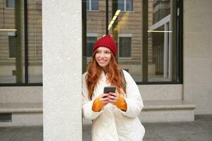 Beautiful redhead girl with smartphone, standing on street, holding mobile phone, using taxi app, connects to public wifi and uses internet, sends a text message photo