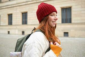 Tourism and travelling. Young redhead woman smiling, tourist walking with backpack around city centre, sightseeing, holds backpack with paper map photo