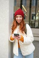 Beautiful redhead girl with smartphone, standing on street, holding mobile phone, using taxi app, connects to public wifi and uses internet, sends a text message photo