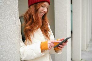 Stylish urban girl using mobile phone app, standing in city, waiting for taxi, looking at smartphone application, texting message photo