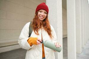 Beautiful redhead woman drinking hot tea or coffee from thermos, female tourist enjoys warm drink, rests during her journey around town photo