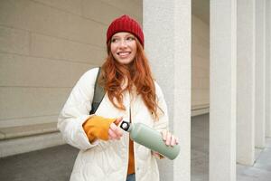 Young happy redhead woman in red hat, drinking from thermos, warming up with hot drink in her flask while walking around city, tourist relaxes with warm refreshemtn photo