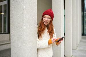 Stylish urban girl using mobile phone app, standing in city, waiting for taxi, looking at smartphone application, texting message photo