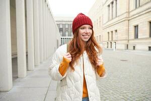 Female tourist in red hat with backpack, sightseeing, explores historical landmarks on her trip around europe, smiling and posing on street photo