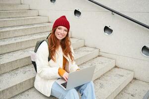 Portrait of young girl traveller, sitting with backpack and map of city, working on laptop, connecting to public wifi and sitting on stairs outdoors, using internet to book hotel room photo