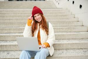 Smiling redhead girl, young woman typing on laptop keyboard, sitting outdoors on stairs with computer, working remote, doing her homework on fresh air photo