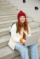 Smiling redhead girl, student sits on stairs outdoors and uses laptop, connects to public wifi in city and works on project, uses internet on computer photo