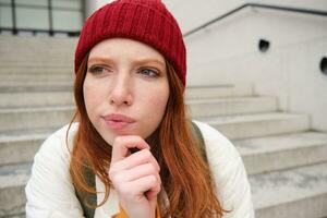 Portrait of redhead girl in red hat, looks thoughtful, touches her chin, frowns and thinks with complicated face expression, sits on stairs outdoors photo