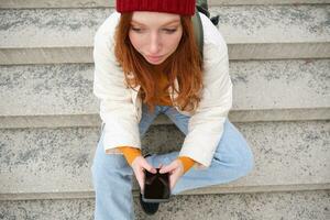 High angle photo of stylish redhead teen girl texts message on phone, uses mobile application while sits outdoors on public stairs