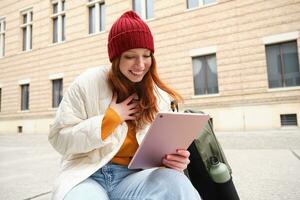 Beautiful redhead woman in red hat, sits with backpack and thermos, using digital tablet outdoors, connects to wifi, texts message, books tickets online photo