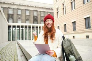 Beautiful redhead woman in red hat, sits with backpack and thermos, using digital tablet outdoors, connects to wifi, texts message, books tickets online photo